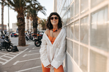 Attractive sporty woman in sunglasse look at camera staying near to wall. Caucasian brunette wearing orange sportscostume and white shirt, Concept of lifestyle