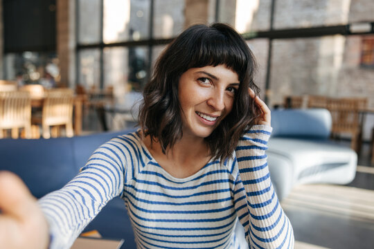Picture of charming european young woman doing selfie with camera and smiling. Staying o the blur background in striped shirt. Lifestyle, positive emotion concept 