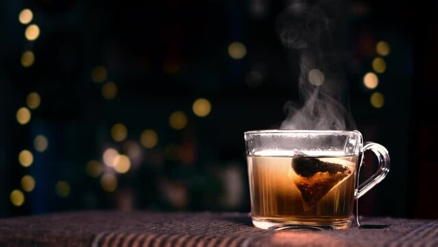 Tea leaves leaching out in hot water. Fresh steaming rooibos or black tea with pyramid or triangular tea bag. Seasonal hot drink. Cozy Christmas mood, glass mug, evening lights, bokeh background. 4K