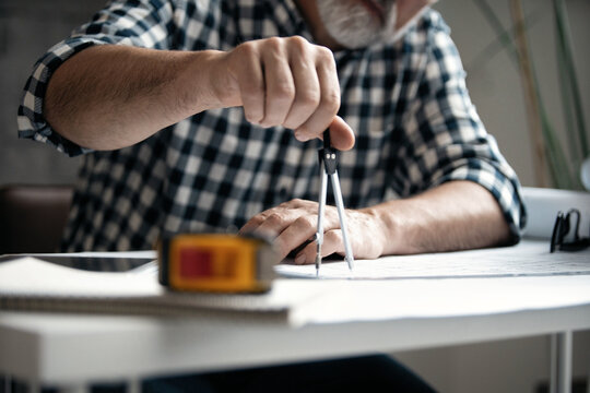 Mature architect working on construction blueprint in modern office. Engineer using divider compass tools on blueprint