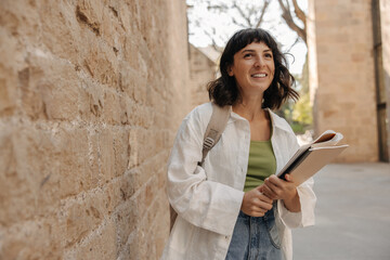 Image of happy student in hands notebooks staying near to wall with backpack. Caucasian brunette woman wearing casual clothes. Concept of lifestyle