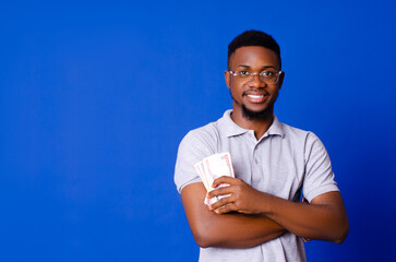 Young handsome African man smiling while holding cash with his arms-crossed, standing on a blue isolated background