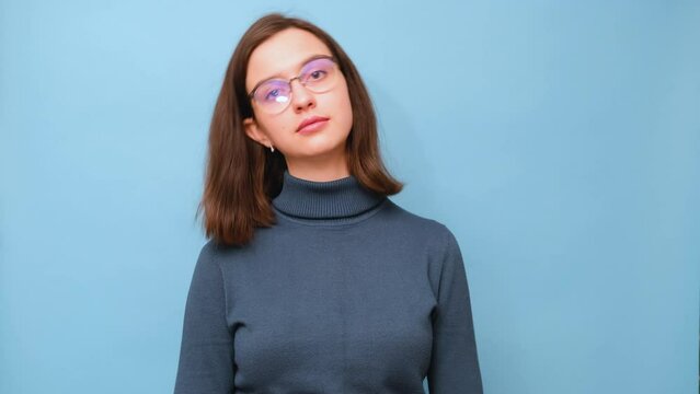 Portrait of a female student in glasses and a sweater waving her head from side to side on a blue background, 4K. Uncertainty. Nonverbal communication. Exercises for the neck. Self-massage