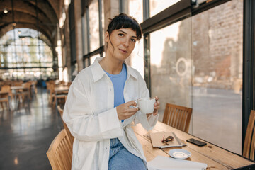 Image of pretty caucasian young woman holding cup coffee staying in cafe. Looking at camera in casual clothes indoors. Rest time concept 