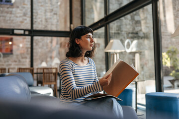 View of beautidul care cute woman looking at window holding notebook. Caucasian brunette in coworking place sitting and studying. Concept of lifestyle - obrazy, fototapety, plakaty