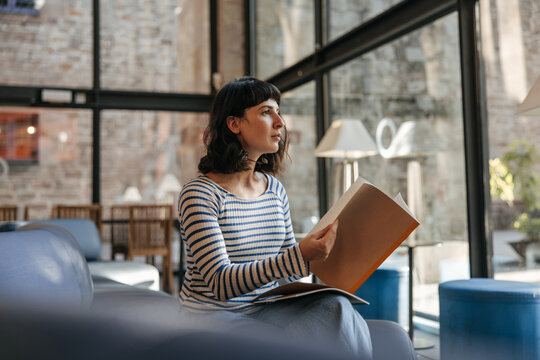 View of beautidul care cute woman looking at window holding notebook. Caucasian brunette in coworking place sitting and studying. Concept of lifestyle