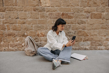 View of pretty dark-haired woman looking at phone and chatting. European with short hair in hands smartphone outdoors. Use technology concept