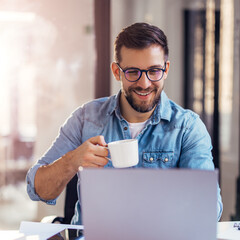 Handsome smiling young man sitting by window in office, drinking coffee while working on laptop.