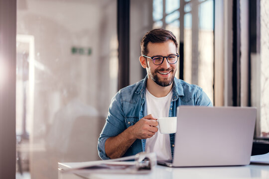 Handsome smiling young man sitting by window in office, drinking coffee while working on laptop.