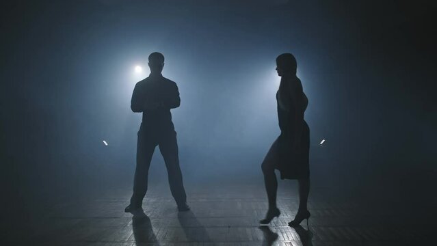 Young woman comes to partner. Young couple dance tango in dark room with smoke and spotlights. Silhouette of dancers in smoky ballroom. Woman legs on heels. Slow motion in 4K, UHD