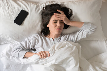 Sad young european woman wakes up, presses her hand to head, suffers from headache, has bad dreams