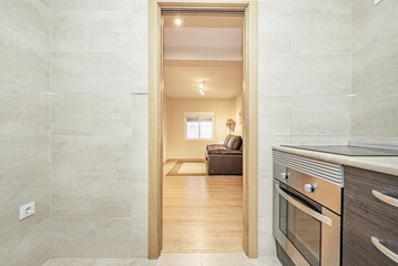 Detail of a kitchen furnished with beige marble tiles and an oak sliding access door overlooking a living room with a leather sofa