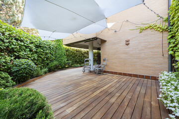 Terrace of a ground floor house with hedges and ornamental plants, slatted acacia hardwood floors twin wooden sunbeds and white awnings - obrazy, fototapety, plakaty