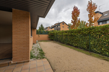 Patio of a semi-detached house with brown stoneware floors and sandy terrain with a bushy hedge separating it from the neighbors