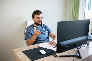 Handsome man smiling with a beard and glasses of сaucasian talks on a video call with colleagues at the workplace with monitors. Programmer, manager, economist, freelancer, having video chat, waving