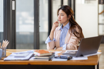 Image of young beautiful brooding Asian woman working with laptop while sitting at laptop in office, thinking of professional plan, project management, considering new business ideas.