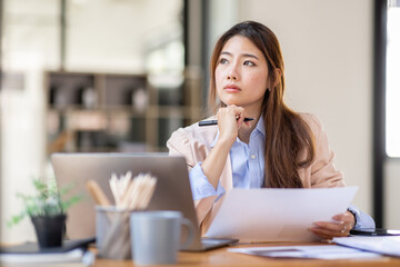 Image of young beautiful brooding Asian woman working with laptop while sitting at laptop in office, thinking of professional plan, project management, considering new business ideas. - Powered by Adobe