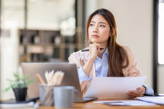 Image of young beautiful brooding Asian woman working with laptop while sitting at laptop in office, thinking of professional plan, project management, considering new business ideas.
