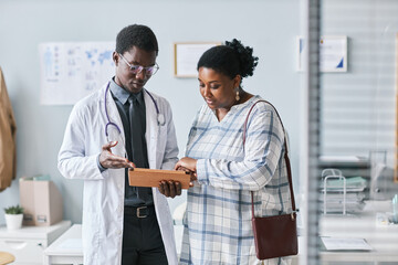 Young black doctor consulting female patient using digital tablet in clinic