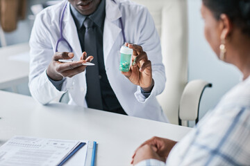 Close up black doctor holding bottle of pills consulting patient in clinic - obrazy, fototapety, plakaty