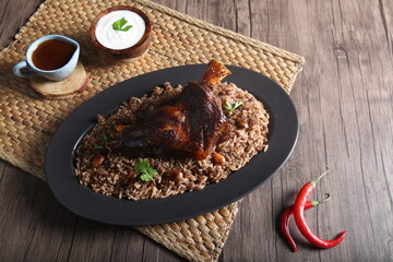 A piece of meat served with brown rice with nuts, brown sauce and yogurt served on a wooden table beside a hot pepper.