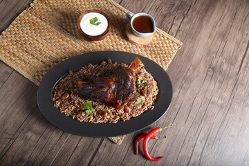 A piece of meat served with brown rice with nuts, brown sauce and yogurt served on a wooden table beside a hot pepper.