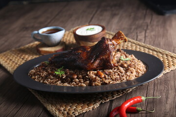 A piece of meat served with brown rice with nuts, brown sauce and yogurt served on a wooden table beside a hot pepper.