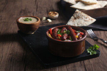 Sausage casserole served with tahini, bread, and garlic on a black tray 
