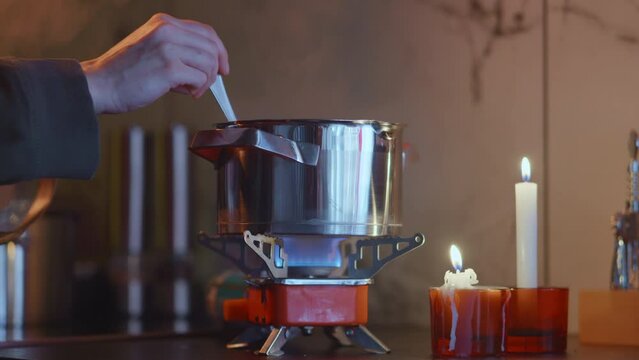 Portable stove usage at home during blackout, cooking on a tourist gas burner, stiring dish in a steel pot. Beauiful domestic background, burning candles. Surviving in Ukraine during war. HQ 4k