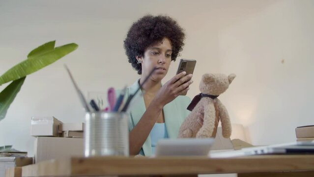 Young Black businesswoman taking closeup shot of teddy bear toy. Short-haired girl taking commercial photo of product for sale with mobile phone in home office. Photography, commerce, startup concept