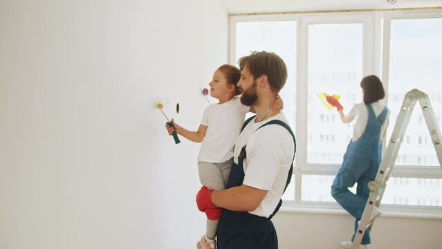 Joyful young Caucasian family painting room. Family time together with their young daughter father, paint a wall with a roller. Dad teaching little adorable daughter to paint wall using roller brush