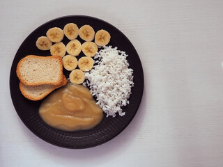 Banana slices, boiled rice, applesauce and toast on a dark plate. Copy space