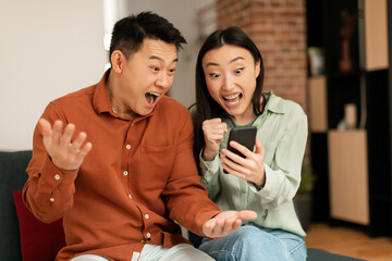 Fototapeta na wymiar Portrait of overjoyed asian couple using smartphone and making winner gesture, shaking fists, sitting on couch at home