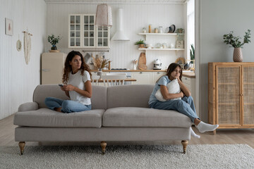 Fototapeta premium Lonely bored teenage girl child sitting on sofa with working online mother, feeling sad, need attention from busy mom. Parent using phone, ignoring daughter. Distracted parenting, internet addiction