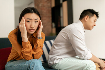 Sad asian wife ignoring upset husband after quarrel, touching head, sitting on sofa in living room, selective focus