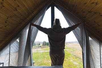 A woman dressed in gray enters an attic room. The ceiling is wooden. The woman opens the curtains. High quality photo