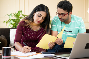 Serious young indian couple calculating expenses or monthly bills in front of laptop while sitting at home - concept of investment, budget and financial planning