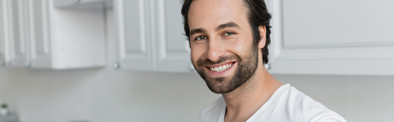 Obraz premium happy and bearded man in white t-shirt looking at camera in kitchen, banner.