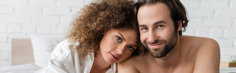 curly woman in silk robe leaning on shirtless boyfriend smiling in bedroom, banner.