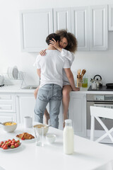 back view of man in jeans seducing curly woman sitting on kitchen worktop.