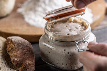 Male hands open a jar with active yeast, flour and fresh bread and pastries in the background