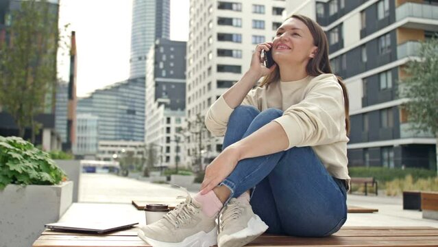 Young cheerful woman talking on a smartphone while sitting on the street of a modern European city.