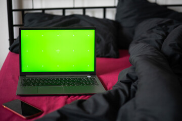 Laptop with green chroma key screen and smartphone on bed with viva magenta bed sheet, black blanket and pillows. Freelancer, blogger, business outsourcing. Home office. Copy space, mockup, template