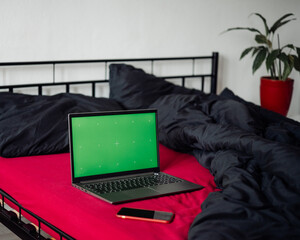 Laptop with green chroma key screen and smartphone on bed with viva magenta bed sheet, black blanket and pillows. Freelancer, blogger, business outsourcing. Home office. Copy space, mockup, template