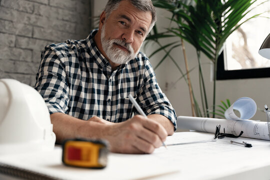 Mature confident smiling architect in casual wear working with blueprint papers and drawings in modern lightful office.