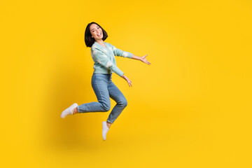 Fototapeta na wymiar Full length photo of cheerful girl dressed blue cardigan jeans jump up arms demonstrate empty space isolated on yellow color background
