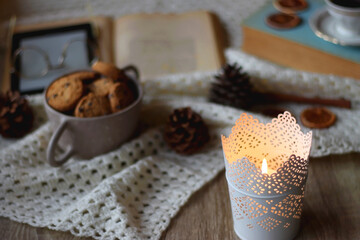 Cup of tea, books, tablet, bowl of cookies, various spices, pine cones and lit candles. Hygge at home. Selective focus.