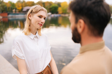 Obraz premium Close up view from back of unrecognizable bearded man to cheerful woman standing holding hands with boyfriend outdoors by beautiful lake at city park, talking and looking at each other smiling.