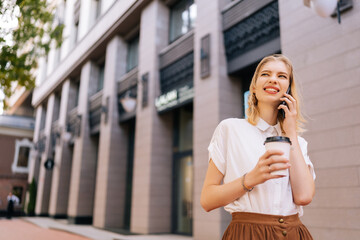 Portrait of young cheerful woman in casual clothes talking on mobile phone while standing on city...
