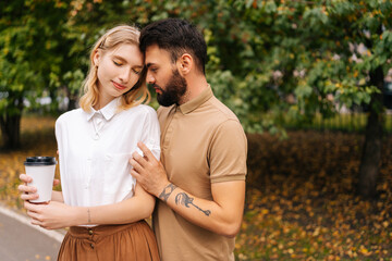 Obraz premium Front view of pretty young couple in love standing embracing with closed eyes at city park on background of trees. Bearded tattooed man and charming woman walking spending time together on summer day.